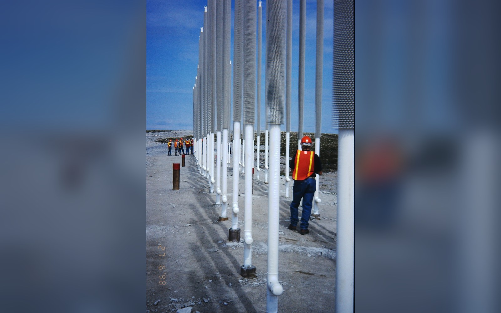 Workers in safety vests inspect rows of tall pipes marking a mineral exploration grid