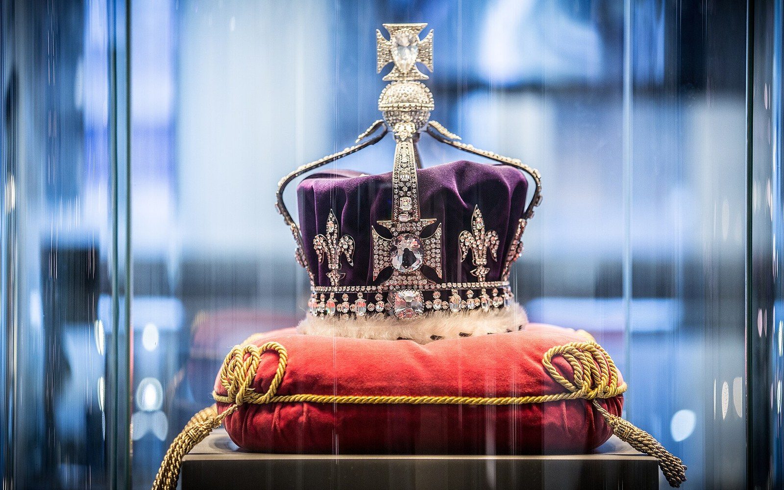 A jewelled crown with a purple velvet cap displayed on a red tasselled cushion in a glass case