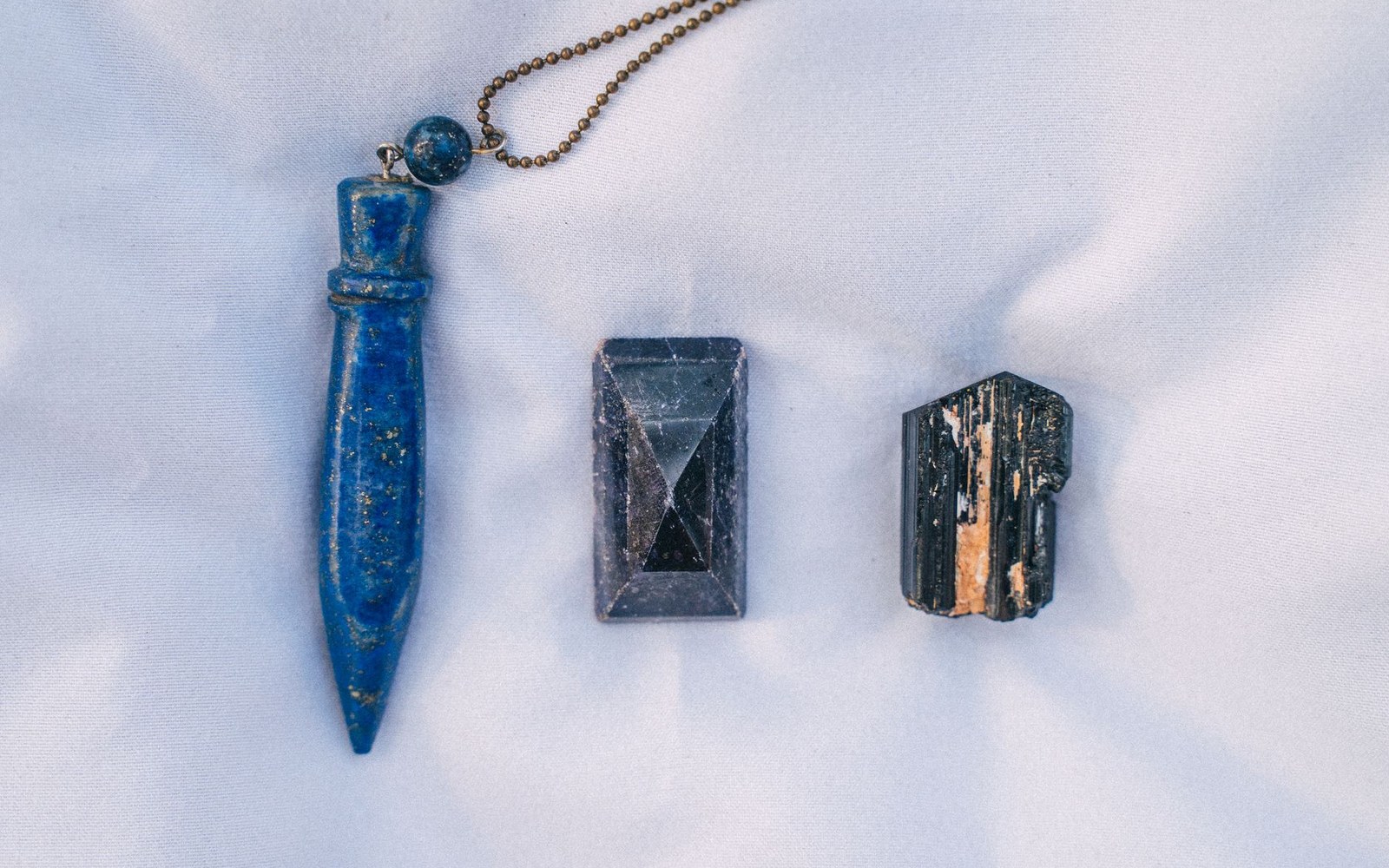 A lapis lazuli pendant on a chain beside a cut lapis piece and raw black tourmaline crystal