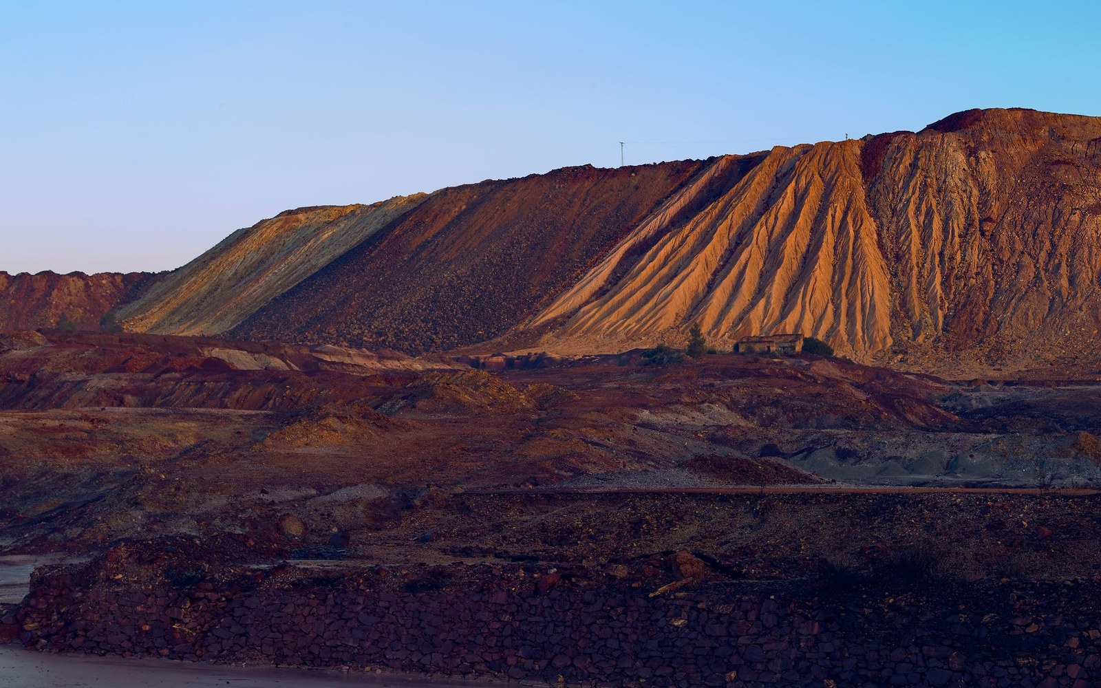 Large rust-colored mine tailings piles rise beside a small building at dusk