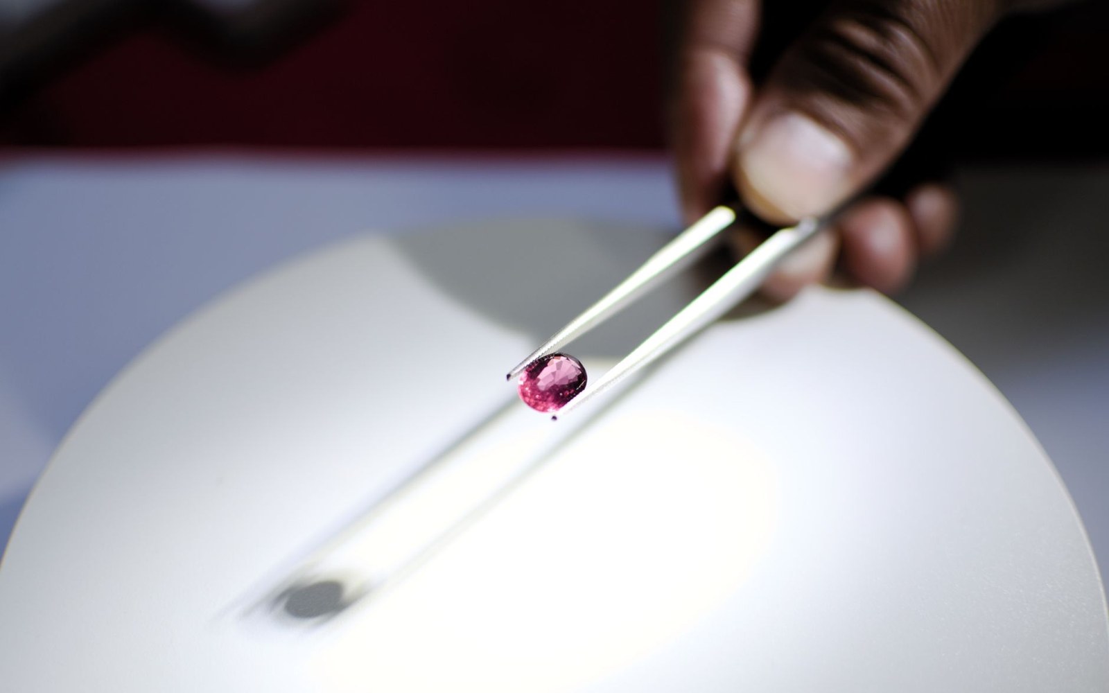 A hand holds a pink gemstone with tweezers above a light box