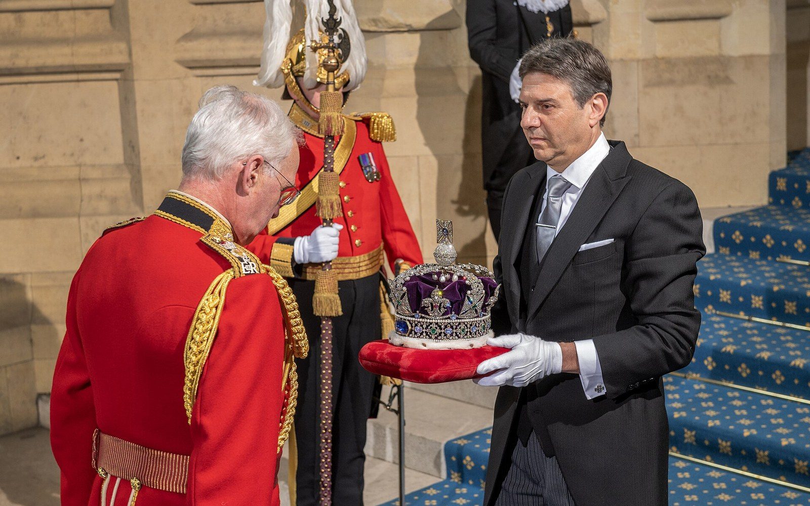 A man in formal morning dress carries the Imperial State Crown on a cushion past uniformed guards